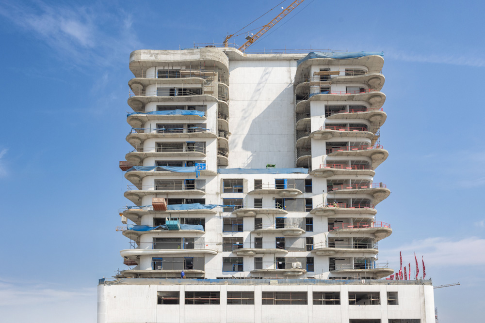 Beach Walk Iii By Imtiaz A partially constructed high-rise building with exposed floors, scaffolding, and blue protective covers against a clear sky.